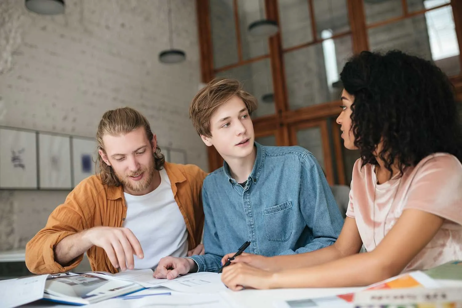 Young cool people working in office. Two boys with blond hair and girl with dark curly hair sitting and studying together in classroom. Students working on new project